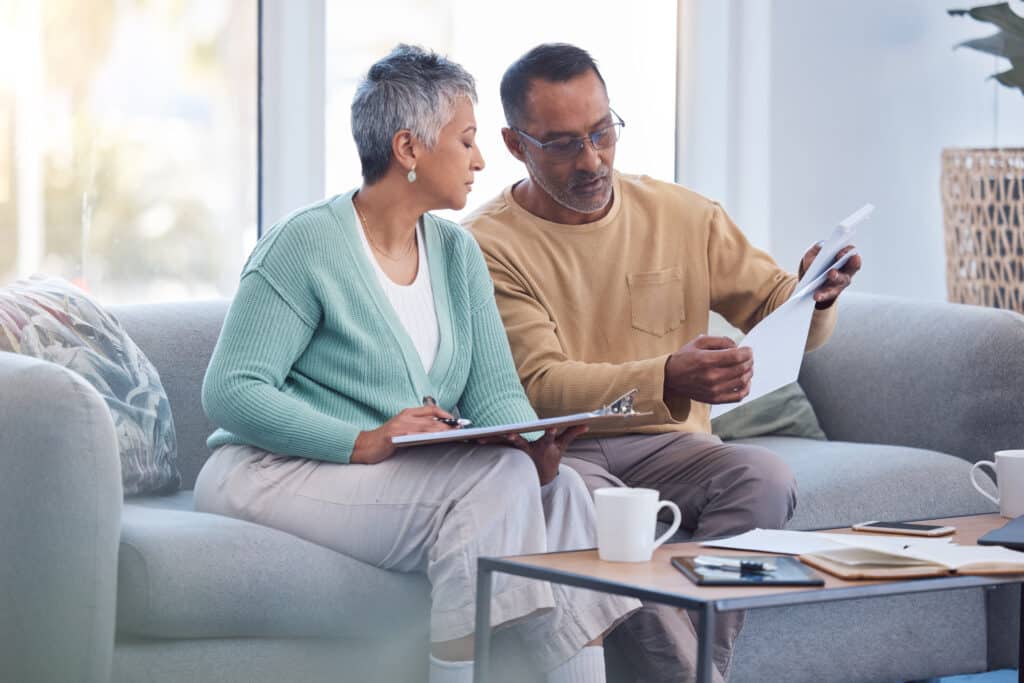 a man and a woman sitting on a couch looking through Medicaid paperwork. They are getting Medicaid application help from the professionals at Brunelle Medicaid