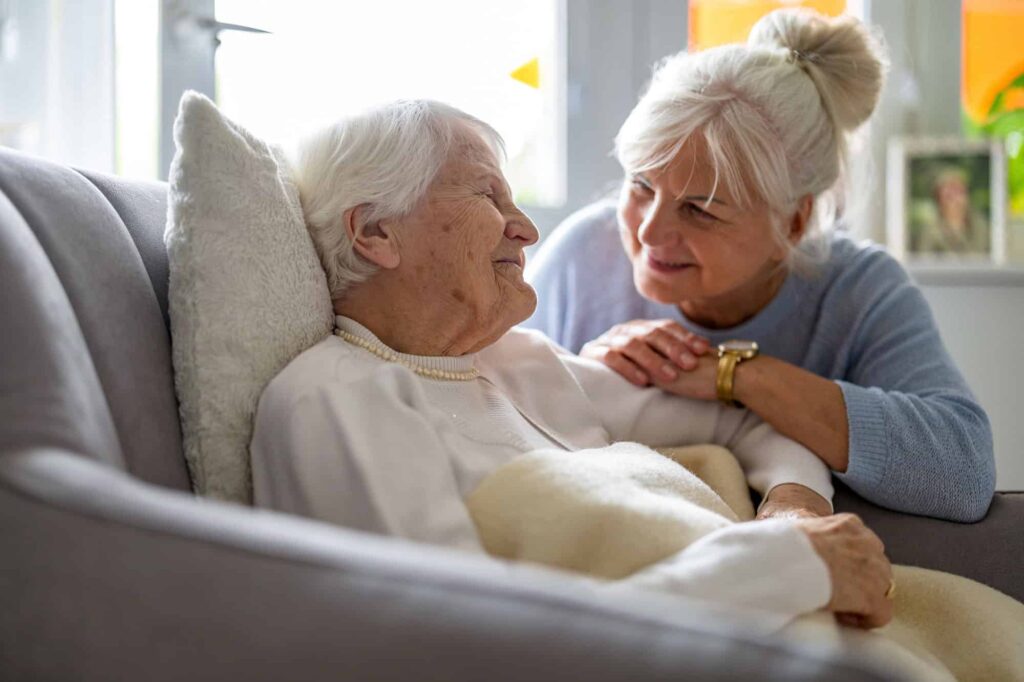 An elderly woman lies on a couch covered with a blanket, smiling at another older woman who sits beside her, gently touching her shoulder. Their caring moment reflects the support families offer when considering who pays for nursing home care.