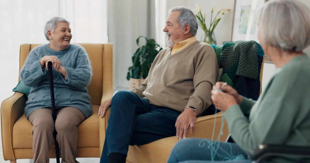 Three older adults sit together indoors, smiling and chatting. One woman holds a cane, another knits, and a man sits between them. Their warm conversation touches on topics like who pays for nursing home care in later life.