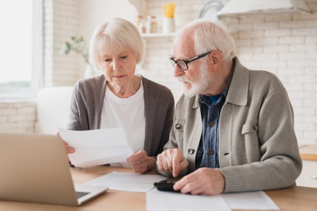 An elderly couple sits at a table with a laptop and documents, looking concerned as they review the income limit for MassHealth for seniors in 2025; the man uses a calculator while the woman holds a sheet of paper.