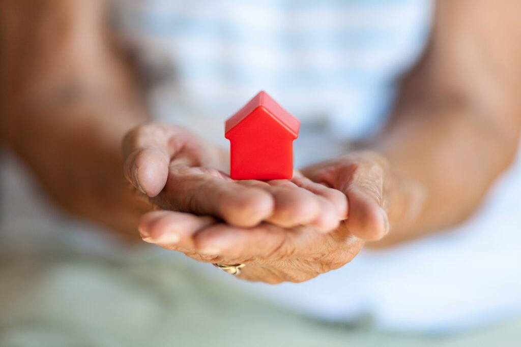 Close-up of an elderly persons hands gently holding a small red toy house, symbolizing home, safety, or real estate. The background is blurred, focusing attention on the hands and house.