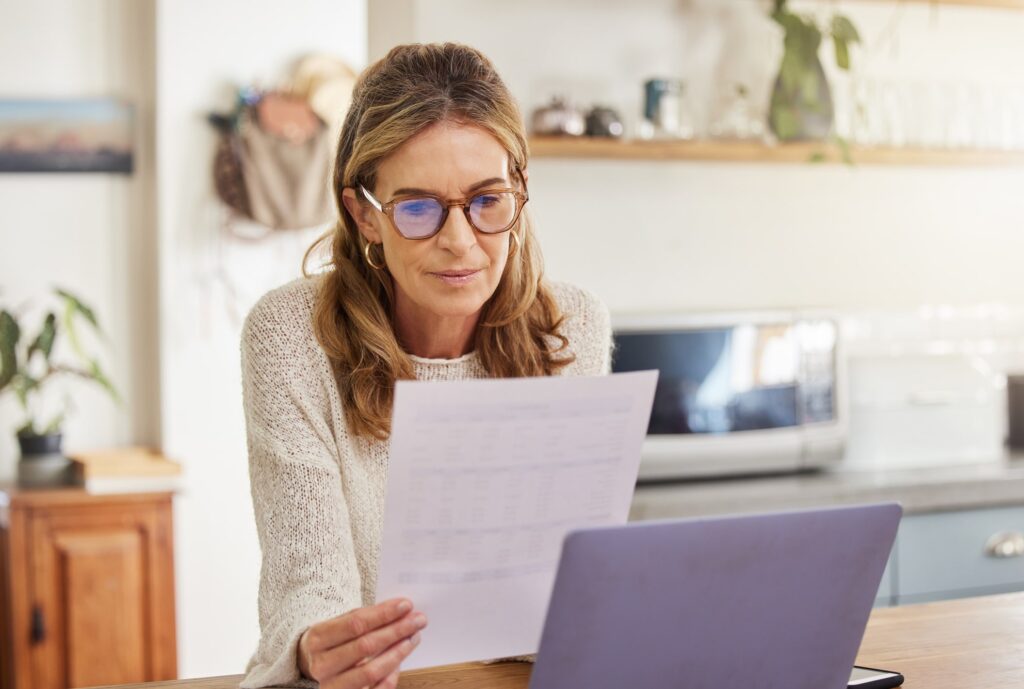 A woman wearing glasses sits at a kitchen table, looking at a sheet of paper while working on a laptop. The kitchen is bright with sunlight and has shelves with various items in the background.