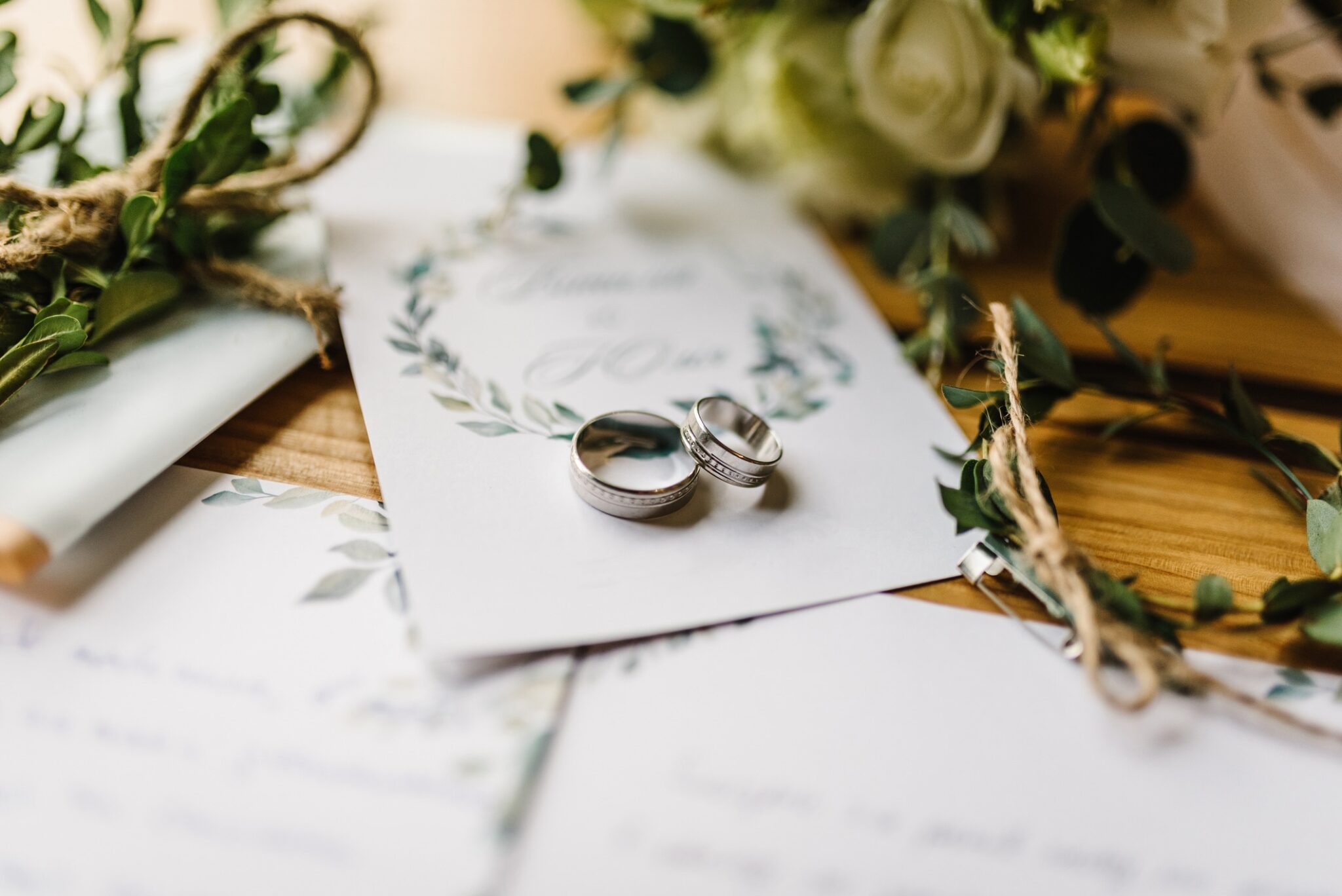 Two silver wedding rings rest on a floral wedding invitation, surrounded by greenery, twine, and handwritten notes on a wooden surface. The scene evokes a romantic and rustic wedding theme.