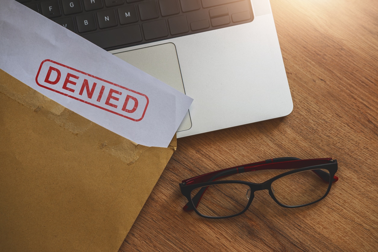 An envelope with a Masshealth application stamped DENIED is placed on a wooden desk next to a laptop keyboard and a pair of eyeglasses. Sunlight shines on the scene.