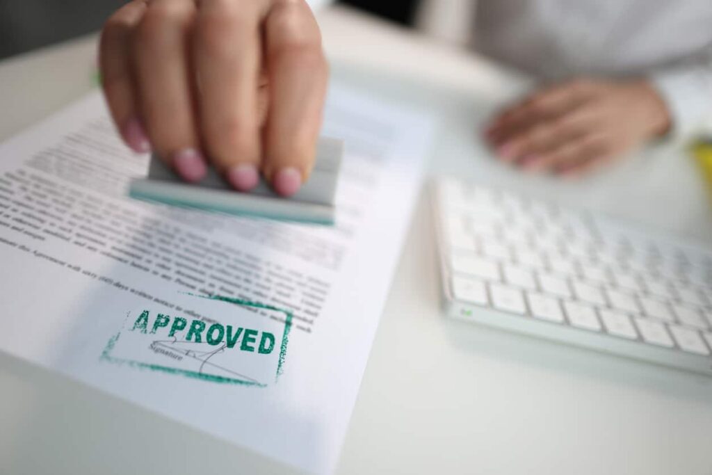 A person stamps a document with a green APPROVED mark beside a computer keyboard on a white desk. The hand and stamp are in focus, while the document text is blurred.