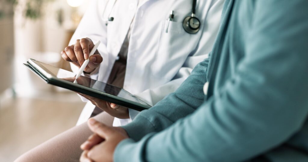 A doctor in a white coat uses a digital tablet with a stylus, to explain how masshealth protects spouses; sitting beside a patient. Both are seated, and only their torsos and hands are visible. The patient’s hands are clasped together in their lap.