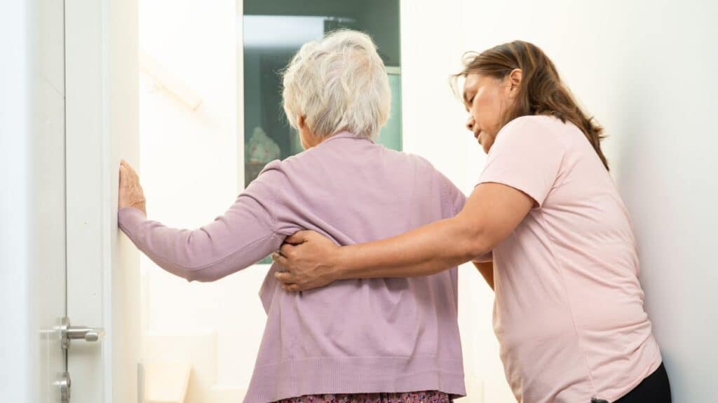 A caregiver supports an elderly woman with gray hair as they walk through a doorway, offering assistance—an everyday moment in the journey toward MassHealth nursing home placement MA, set in a brightly lit hallway and seen from behind.