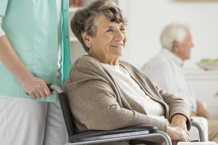 Smiling Elderly Woman In A Brown Sweater Sits In A Wheelchair, With A Caregiver Standing Beside Her. Another Older Man Is Seated In The Background In This Bright, Indoor MassHealth Nursing Home Placement MA Setting.