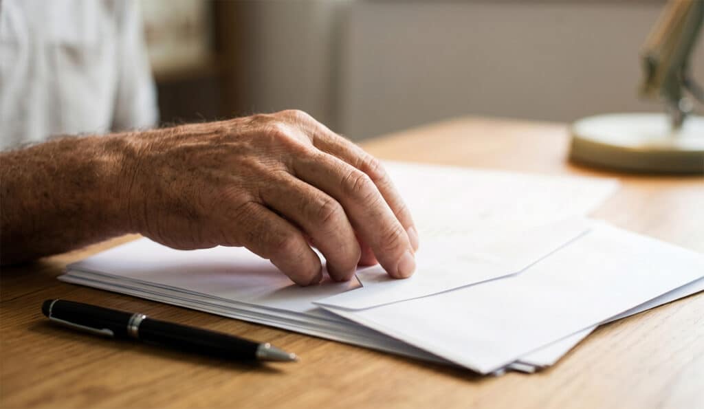 A person’s hand sorting through a stack of papers on a wooden desk, with a pen resting nearby. The scene is well-lit and suggests paperwork or document review.